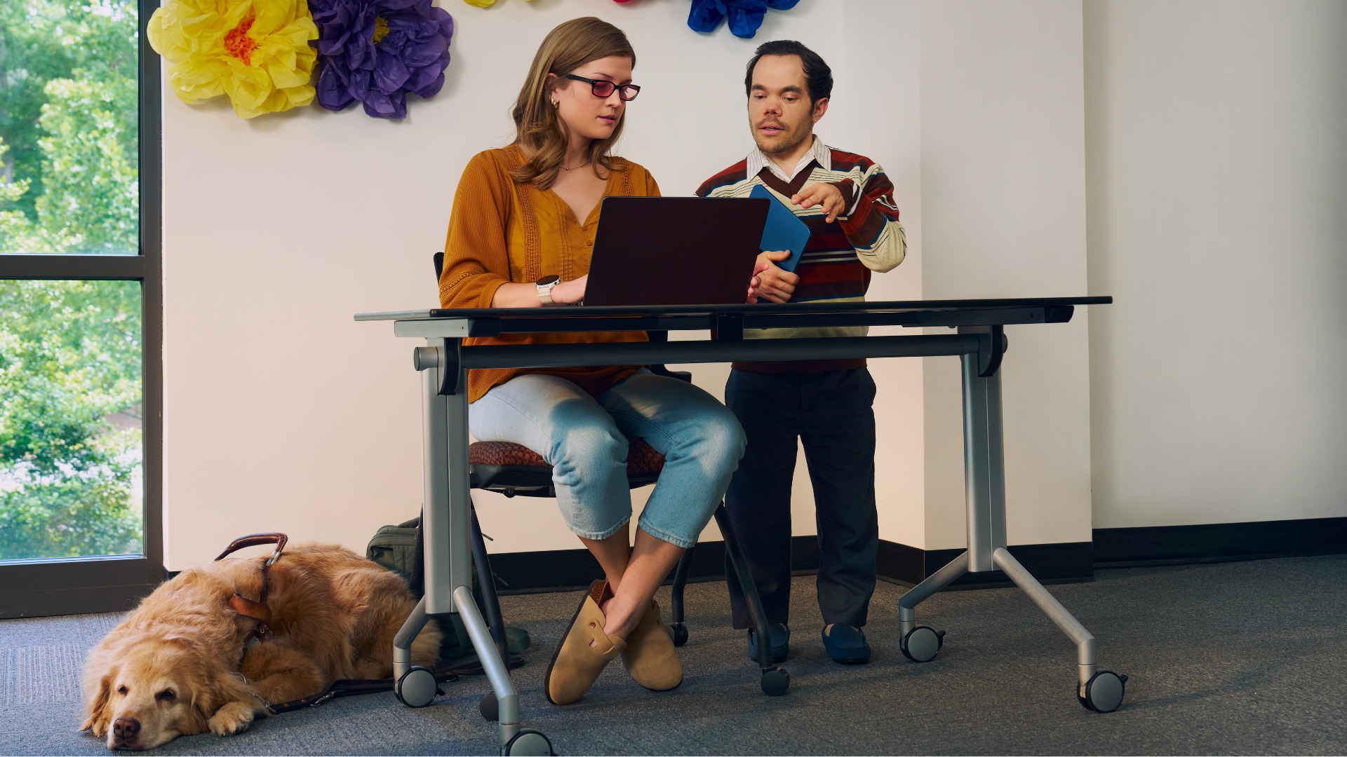 A blind, white woman wearing glasses with red lenses sitting at a lecture table, chats with her professor, a white man with medium brown hair and light brown eyes who has dwarfism, who is standing next to her. They are both looking intently at the student's laptop. A golden retriever service dog wearing a guide harness lays on the floor next to her. The student has dark blonde hair and is wearing an orange blouse and jean capris. A large flower sculpture hangs on the wall behind the lecture table. Next to the sculpture is a large window showing many large trees.